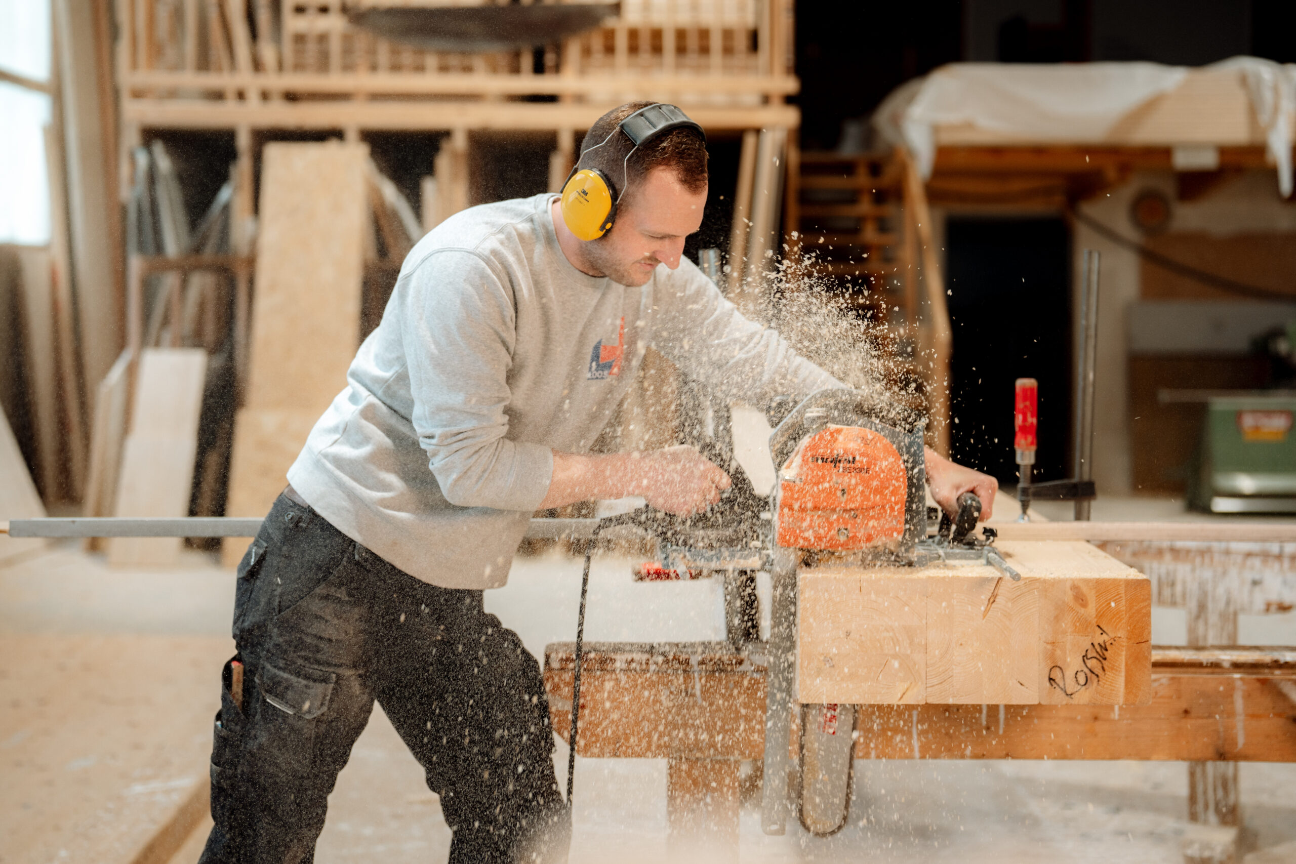 Ein Mann mit Gehörschutz und grauem Sweatshirt schneidet in einer Werkstatt mit einer Elektrosäge einen Holzbalken durch, wobei ein Sprühnebel aus Sägemehl entsteht. Verschiedene Holzbearbeitungswerkzeuge und -materialien sind zu sehen - perfekt für Webdesign oder SEO Agentur Inspiration. fotograf-webdesigner-seo-agentur-hochzeitsfotograf-abgelichtet