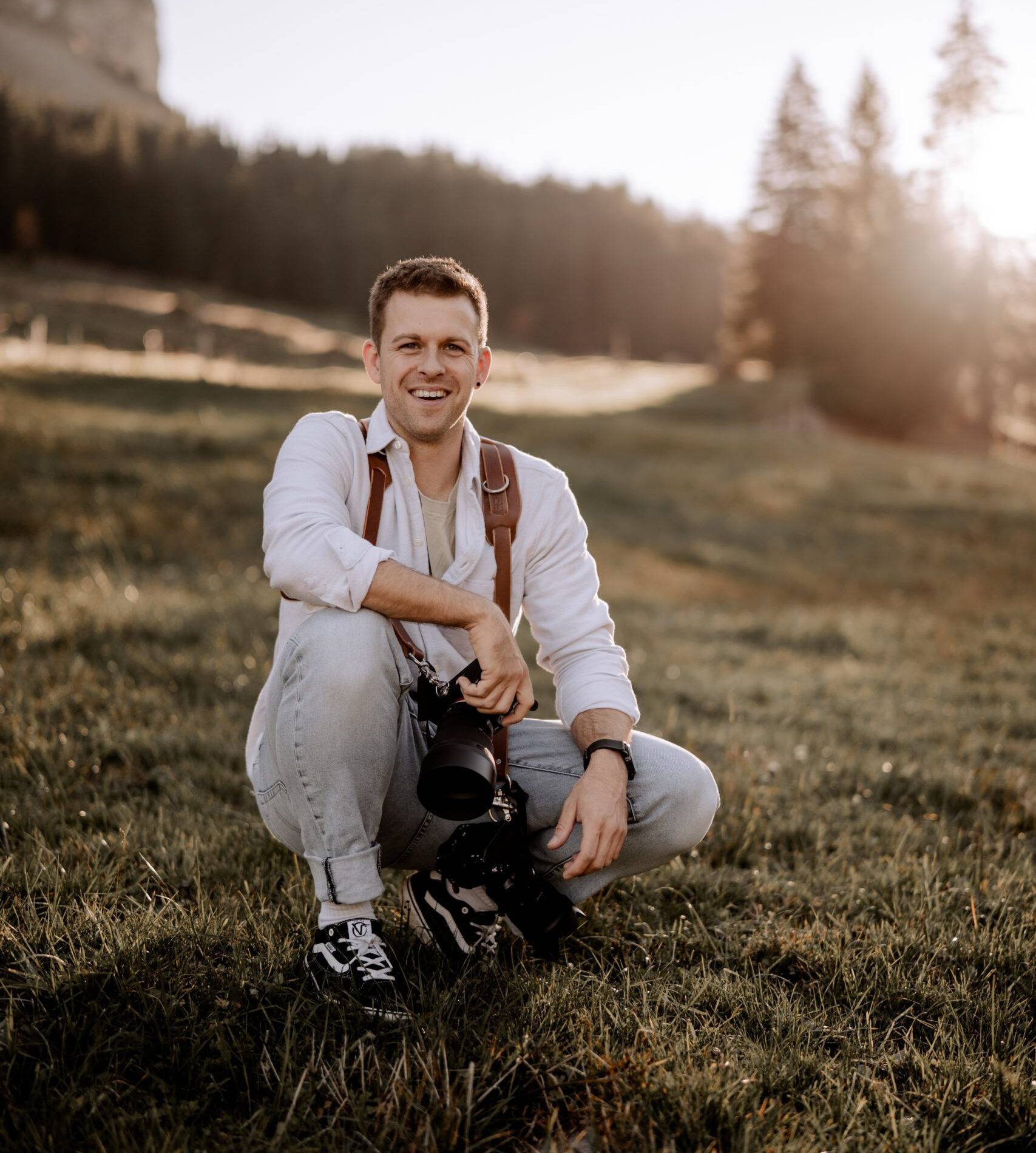 Ein Fotograf in einem weißen Hemd und hellen Jeans hockt im Gras in einer sonnenbeschienenen ländlichen Landschaft, hält eine Kamera und lächelt. Im Hintergrund sind Bäume und ein Berghang zu sehen. fotograf-webdesigner-seo-agentur-hochzeitsfotograf-abgelichtet