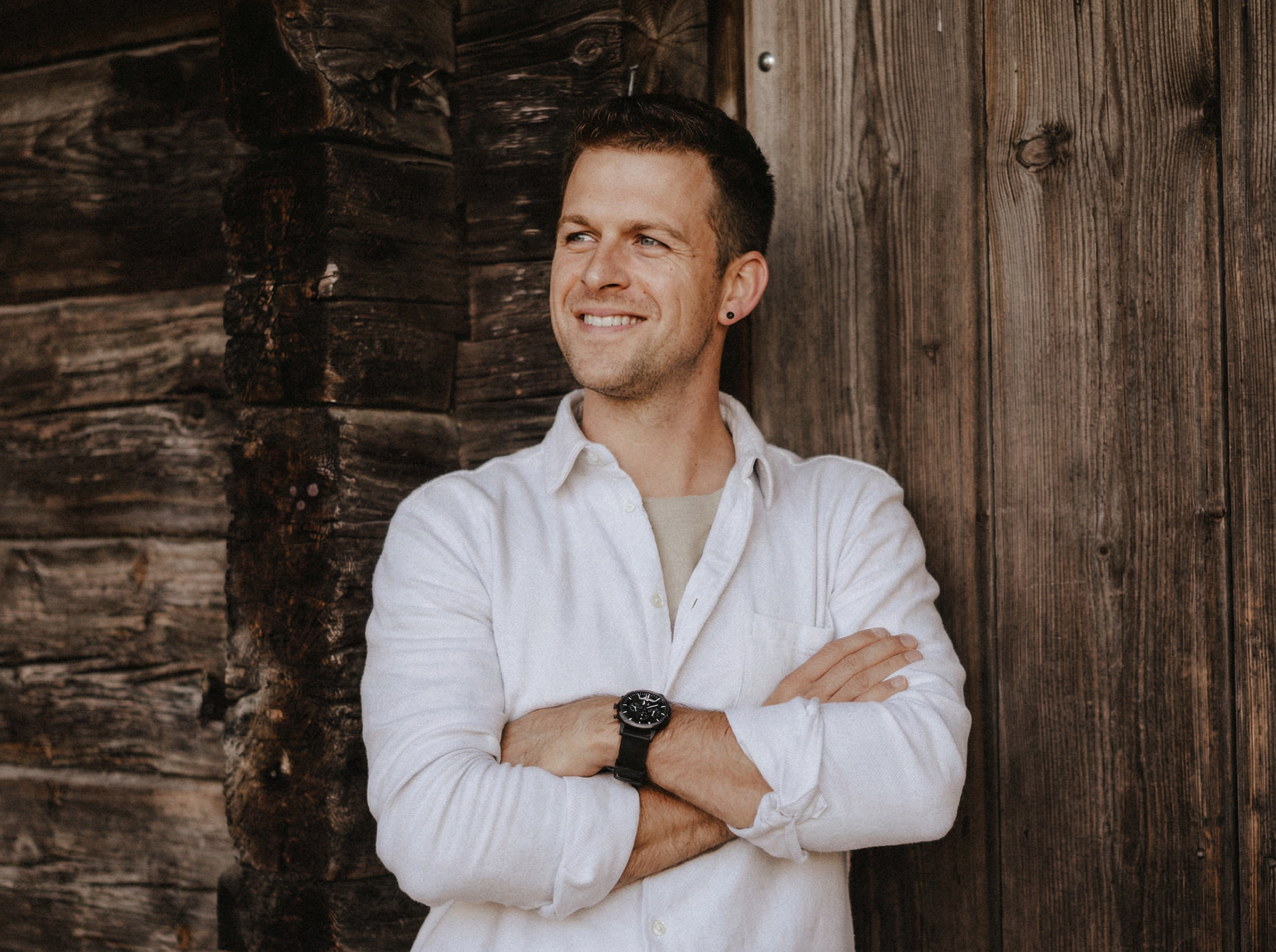 Ein Mann in einem weißen Hemd steht mit verschränkten Armen da, lächelt und schaut zur Seite, während ein Fotograf ihn vor einer rustikalen Holzwand festhält. fotograf-webdesigner-seo-agentur-hochzeitsfotograf-abgelichtet