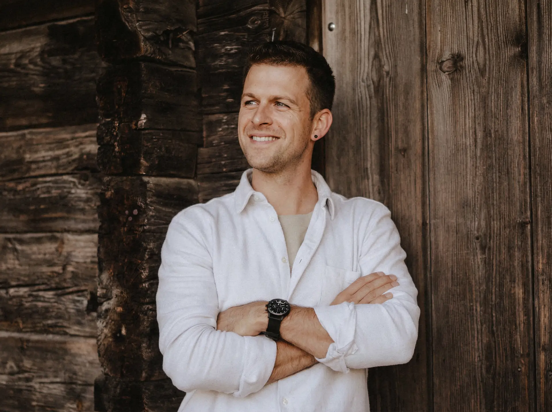 Ein Mann in einem weißen Hemd steht mit verschränkten Armen da, lächelt und schaut zur Seite, während ein Fotograf ihn vor einer rustikalen Holzwand festhält. fotograf-webdesigner-seo-agentur-hochzeitsfotograf-abgelichtet