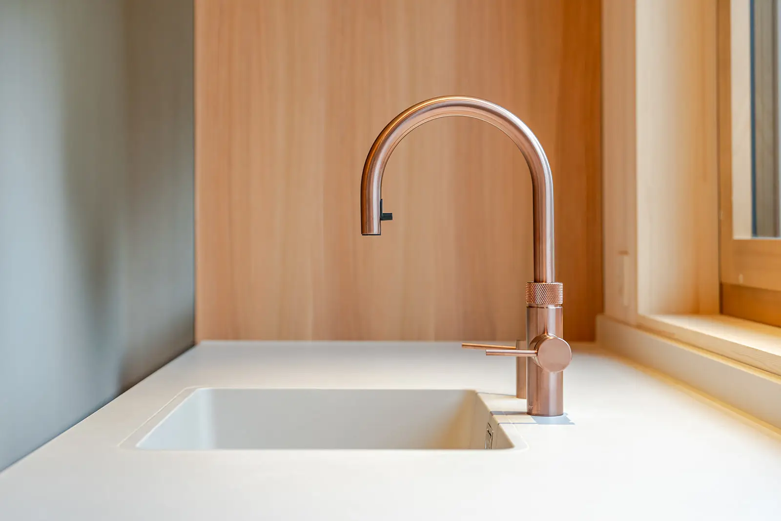 Eine moderne Küchenspüle mit weißem Becken und einem eleganten, geschwungenen Wasserhahn aus Kupfer, vor einer hellen Holzleiste und neben einem Fenster, durch das natürliches Licht einfällt. fotograf-webdesigner-seo-agentur-hochzeitsfotograf-abgelichtet